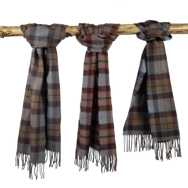 Three Tartan Scarves - OUTLANDER Lambswool in different color patterns (blue-gray, red-gray, brown-blue) are hanging on a wooden rod. Each scarf has fringed ends.