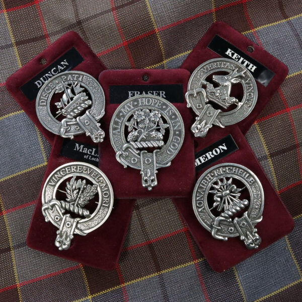 Five Pewter Clan Crest Cap Badge/Brooches displayed on red velvet backings, each featuring detailed crests and names, arranged on a plaid fabric background.