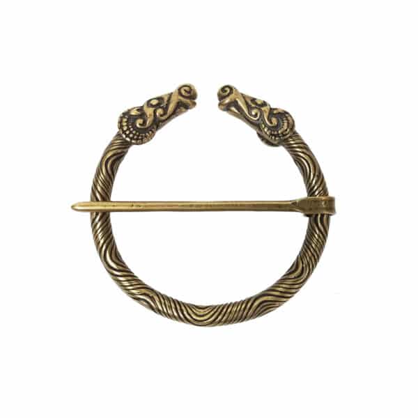 The Ram Penannular Brooch is a circular accessory featuring two ornate dragon heads at the ends and a pin crossing the middle. Reminiscent of ancient designs, this brooch appears to be made of a brass-like metal with intricate patterns on the band.