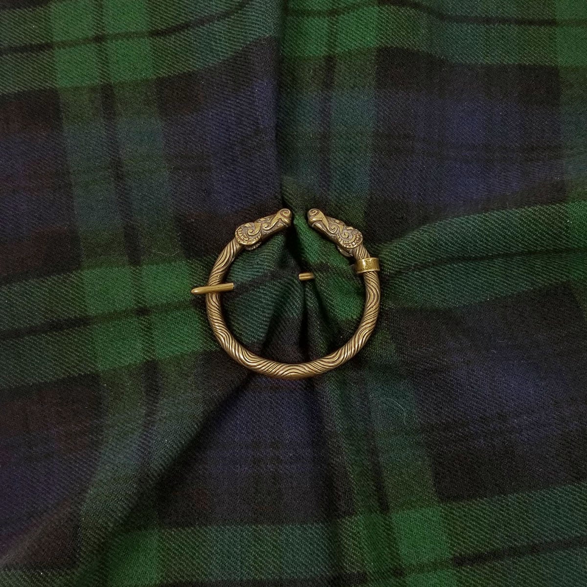 A Ram Penannular Brooch with intricate detailing rests on a green and black tartan fabric.