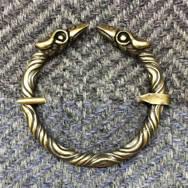 A Raven Penannular Brooch, featuring a brass bracelet with a twisted design and bird heads at both ends, is placed on a textured fabric background.