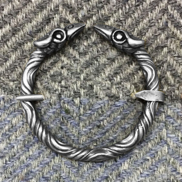A Raven Penannular Brooch, featuring silver twisted arms with two raven-shaped endpoints, is displayed on a herringbone-patterned fabric background.