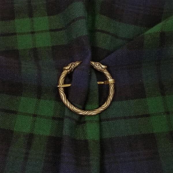 A Raven Penannular Brooch with intertwined snake heads is used to hold together a piece of dark green and navy blue tartan fabric, evoking the mystique of a raven's midnight flight.