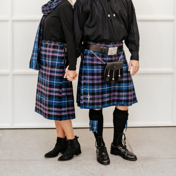 Two individuals holding hands, dressed in matching plaid kilts and black tops, with one also wearing a blue plaid scarf and the Celtic Embossed Quality Leather Kilt Belt. They are standing indoors against a white wall.