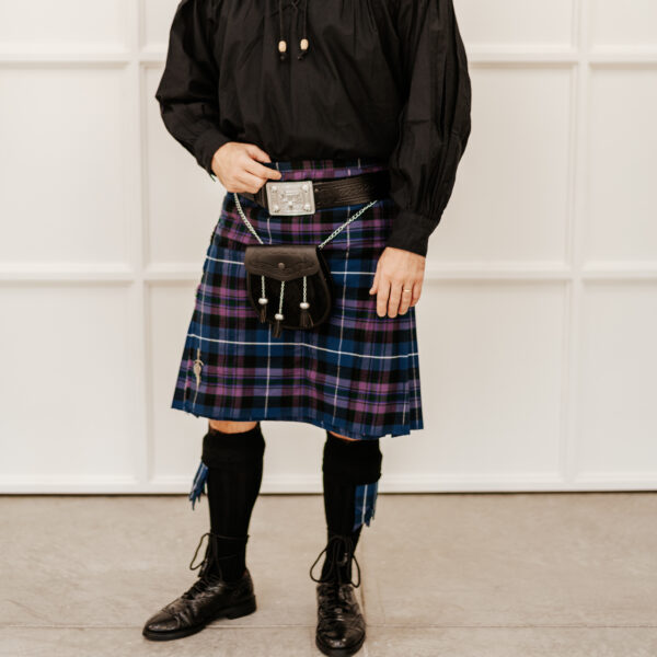 A man wearing a traditional Scottish kilt ensemble, including a black shirt, black sporran, black shoes, and dark blue tartan-patterned kilt, stands proudly against a white panel backdrop. He completes his attire with the Celtic Embossed Quality Leather Kilt Belt.