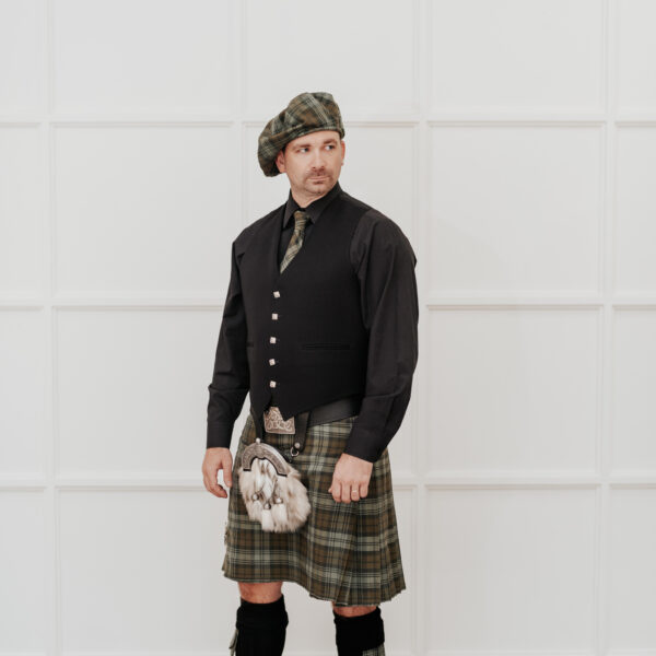 A man in green tartan attire, including a kilt, sporran, and beret, paired with the Quality Pebble Grain Leather Kilt Belt, stands against a white paneled background looking to the side.