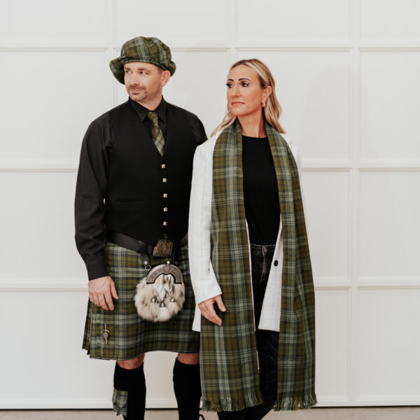 A man and a woman wearing matching tartan outfits stand in front of a white paneled wall. The man wears a kilt, paired with a Quality Pebble Grain Leather Kilt Belt, hat, and vest, while the woman wears a long scarf and a white coat.