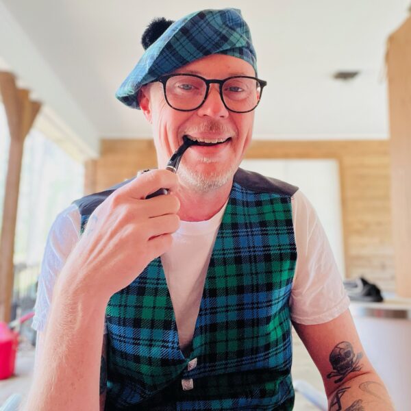 A man wearing glasses and a blue-green tartan outfit, including a matching beret and Men's Tartan Vest Spring Weight 8 oz. wool, smokes a pipe indoors. A table and wooden wall are in the background.