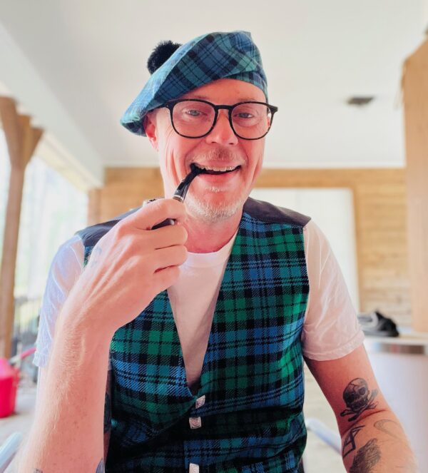 A man wearing glasses and a blue-green tartan outfit, including a matching beret and Men's Tartan Vest Spring Weight 8 oz. wool, smokes a pipe indoors. A table and wooden wall are in the background.