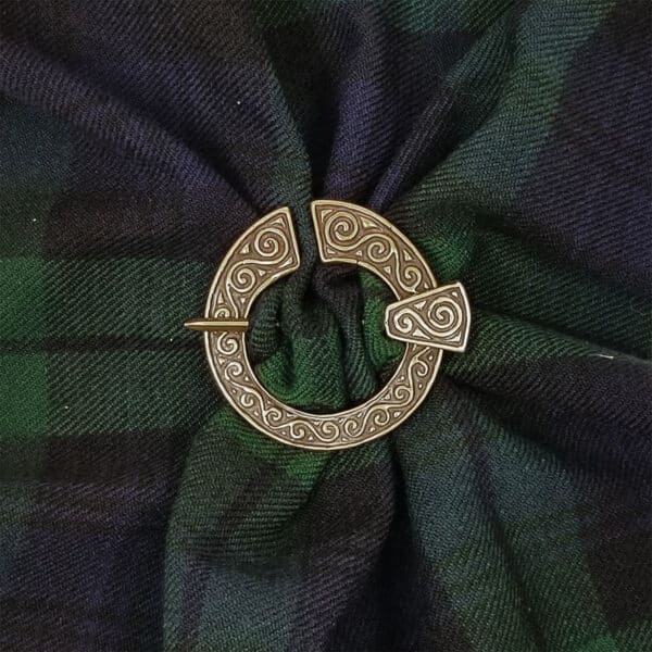 A green and blue tartan fabric, evocative of Dal Riata heritage, is gathered and fastened with the Dal Riata Brooch, featuring a bronze spiral-patterned design.