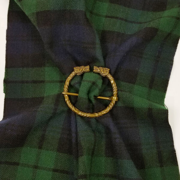 A Wolf Penannular Brooch, featuring a circular design with two gold-tone wolf heads, is pinned to green and blue tartan fabric.