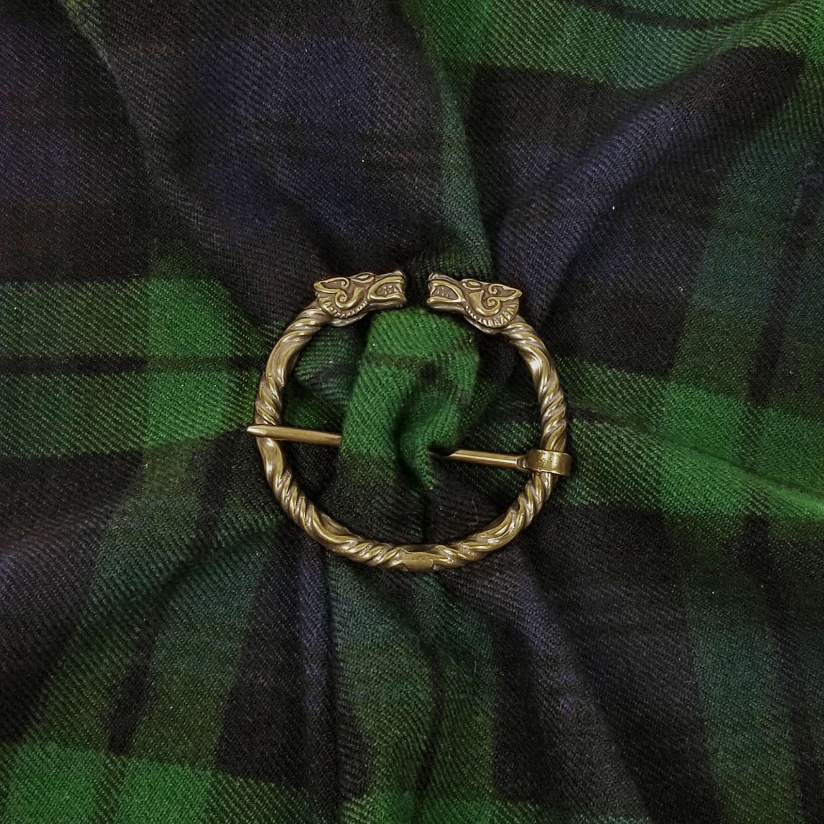 A Wolf Penannular Brooch, featuring a circular design with two gold-tone wolf heads, is pinned to green and blue tartan fabric.