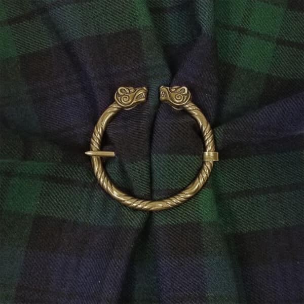 A Bear Penannular Brooch made of brass features serpentine heads clasping a fabric in a green and navy plaid pattern.