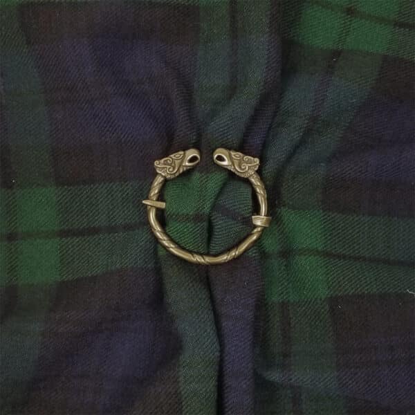 The Griffin Penannular Brooch, featuring two ornate animal heads resembling griffins, holds together a piece of dark green and navy plaid fabric.