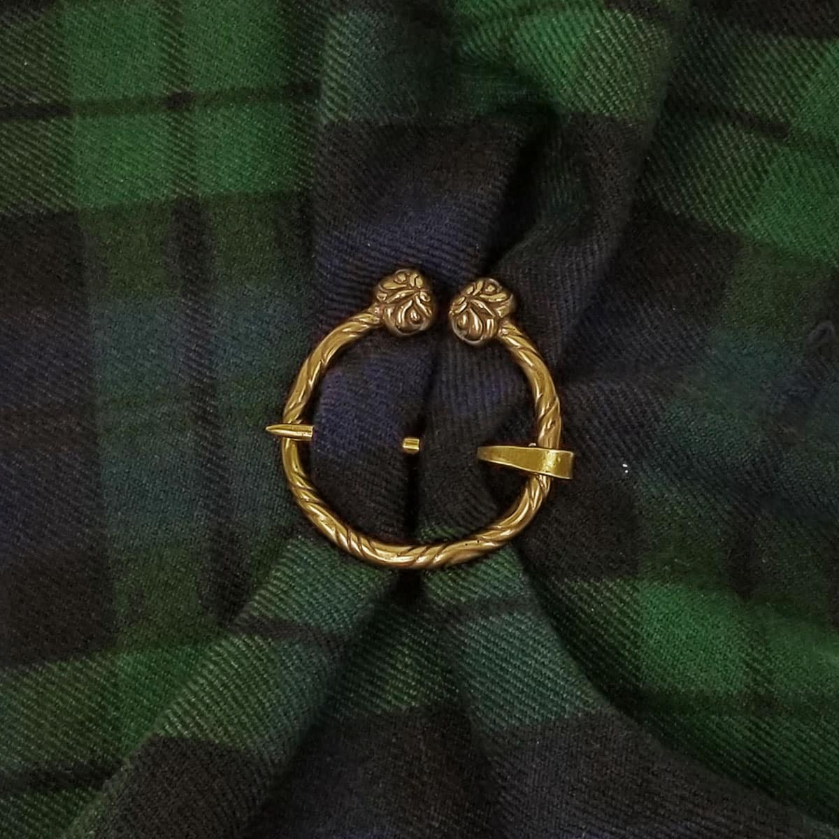 An Owl Penannular Brooch is pinned to a piece of dark green and black tartan fabric.