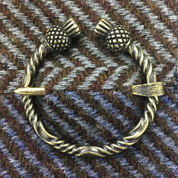 A Thistle Penannular Brooch with a bronze twisted design and decorative spherical ends rests on a tweed fabric background with a herringbone pattern.