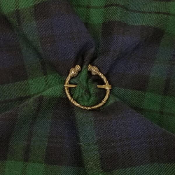 A black and green tartan cloth is gathered and fastened with the Thistle Penannular Brooch, which features intricate gold thistle designs.