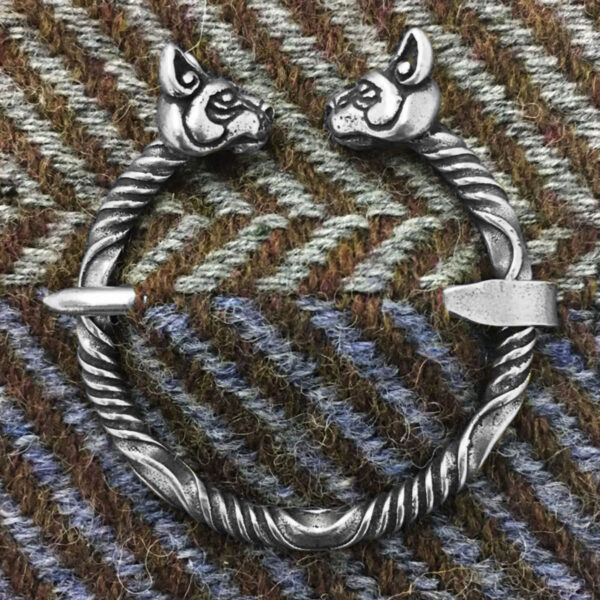 The Cat Penannular Brooch showcases a circular, twisted design with two animal head motifs at the ends, all set against a woven fabric background.