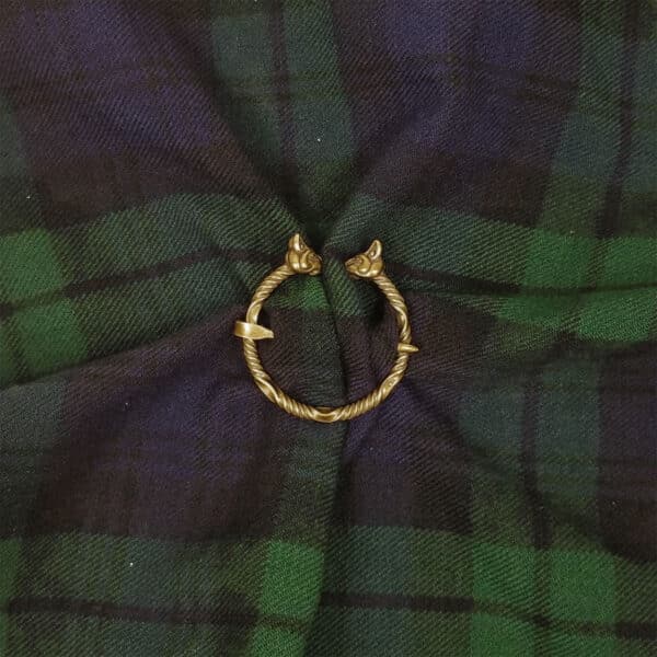 A Cat Penannular Brooch, featuring animal heads on each end, is placed on a crumpled dark green and black tartan fabric.