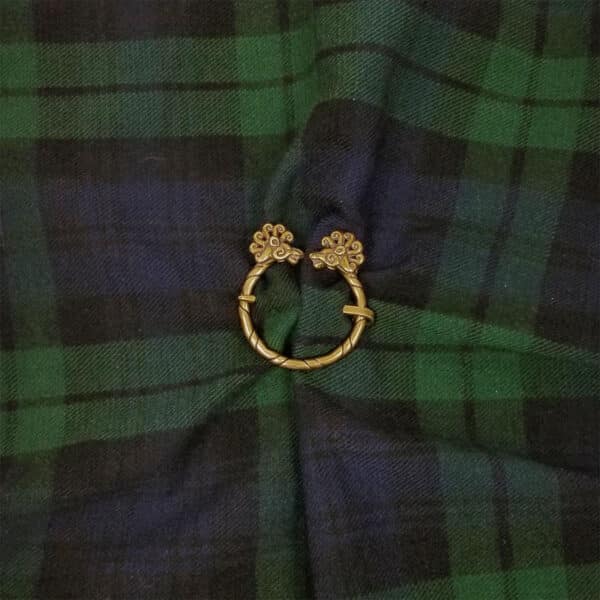 A Lion Penannular Brooch, featuring intricate spiral designs in bronze, lies on a green and blue tartan fabric background.