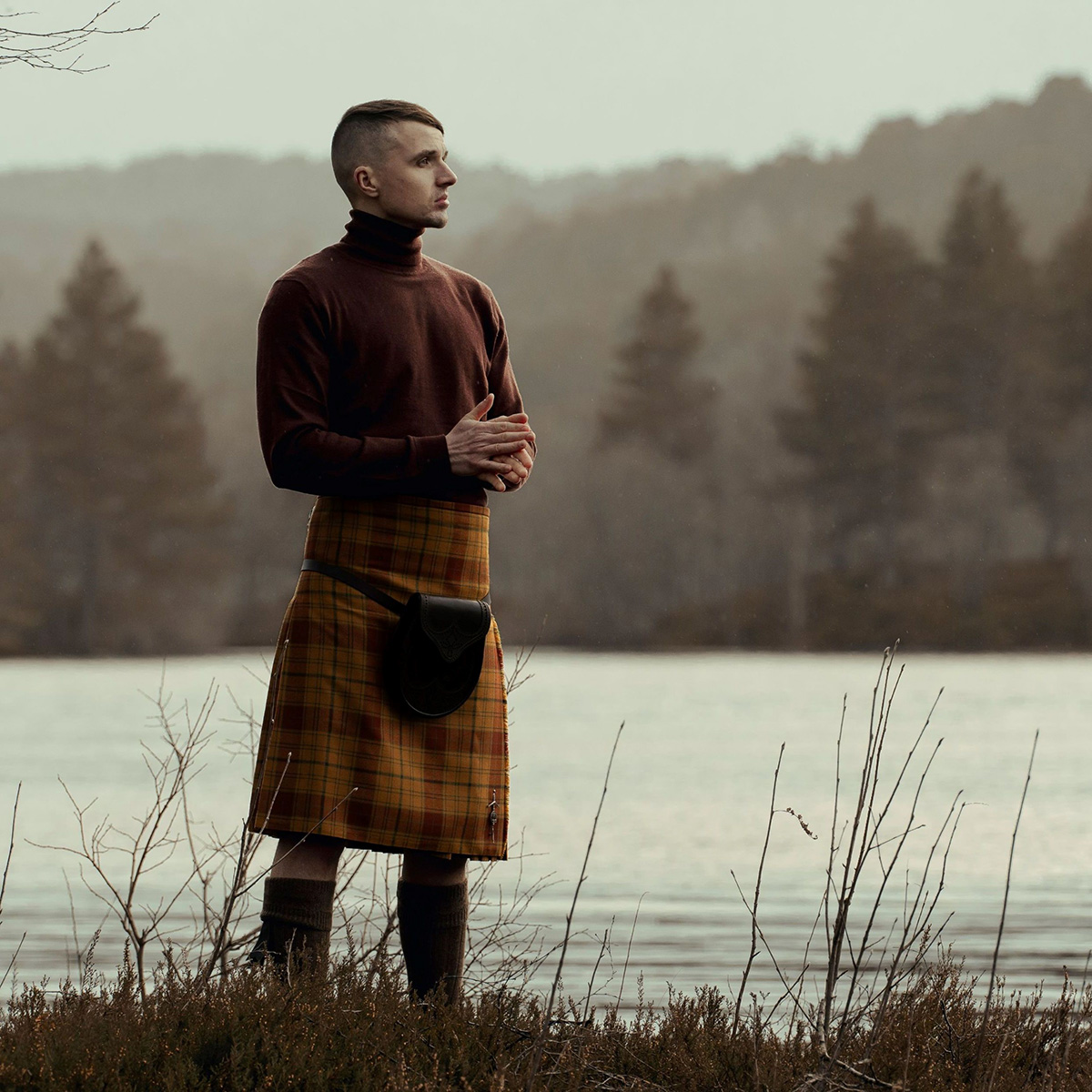 A person stands by a lake wearing a brown long-sleeve top and a 9 Yard Medium Weight 13oz Premium Wool Formal Kilt, with their hands clasped. Trees and misty hills are in the background.