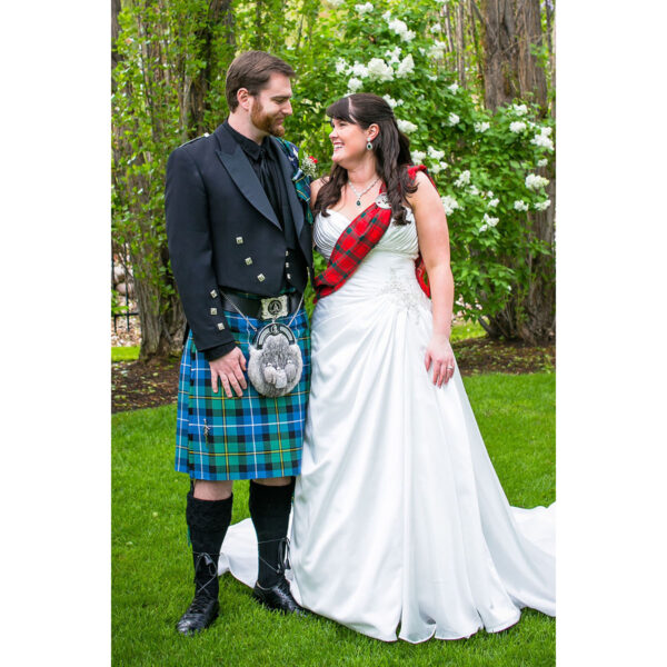 A couple in wedding attire stands on grass. The groom, in a 9 Yard Medium Weight 13oz Premium Wool Formal Kilt with a sporran, and the bride, in a white gown adorned with a tartan sash. Trees and bushes form the background.