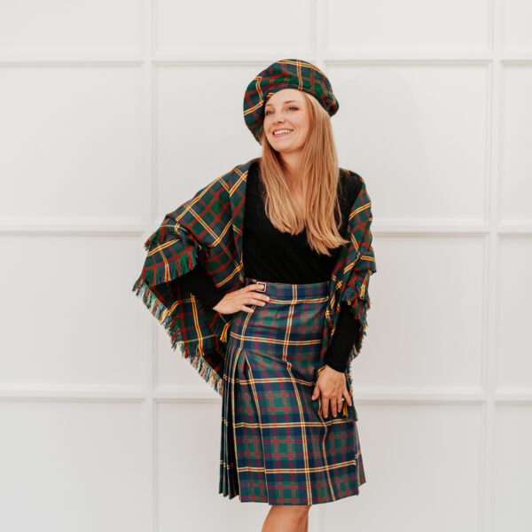 A woman stands smiling with one hand on her hip, wearing a black long-sleeved top, a plaid skirt, a plaid beret, and the Matching Shawl.