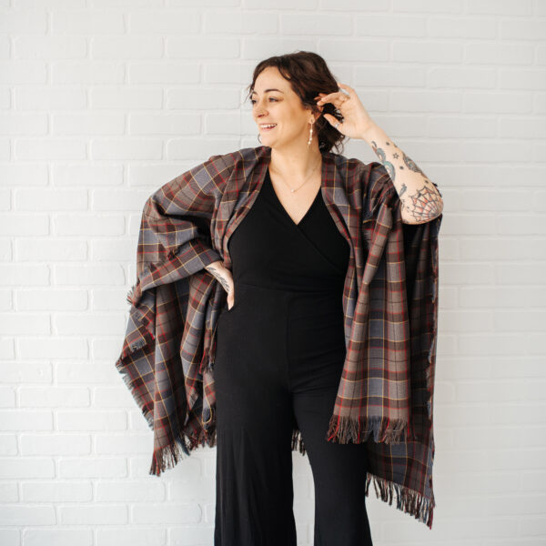 Person wearing a Matching OUTLANDER P/V Serape and black outfit, standing against a white brick wall, smiling and touching their hair.