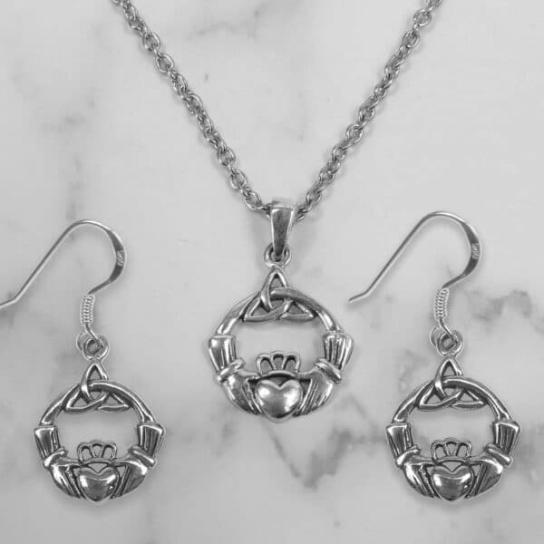 Silver jewelry set on a marble background, consisting of a pendant necklace and matching sterling silver Triquetra Claddagh earrings, each featuring a heart, crown, and hands motif.