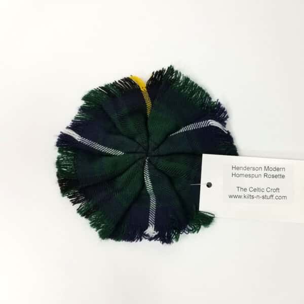 A fabric rosette in green, black, and white tartan with a label reading "Leatherneck Wool-Blend Tartan Rosette, The Celtic Croft, www.kilts-n-stuff.com.
