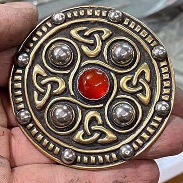 A person is holding a Jeweled Saxon Disc Brooch with a red stone.