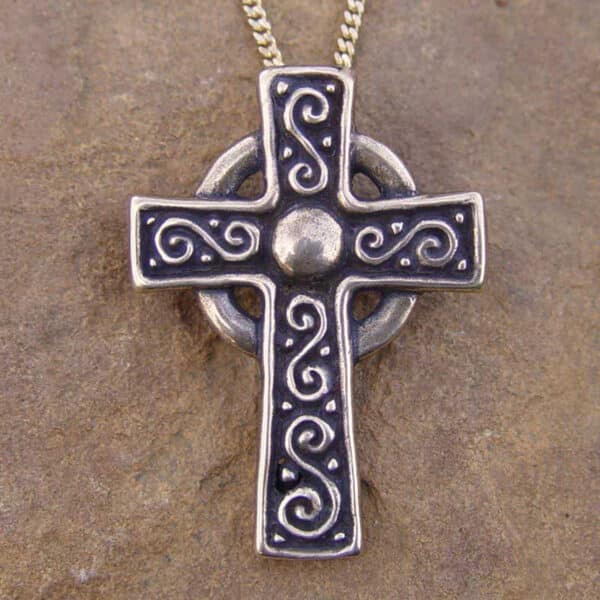 The Celtic Cross Pendant, crafted in silver and featuring exquisite swirl patterns, hangs elegantly from a chain against a stone background. This stunning piece of jewelry embodies the timeless artistry and tradition of Celtic design.