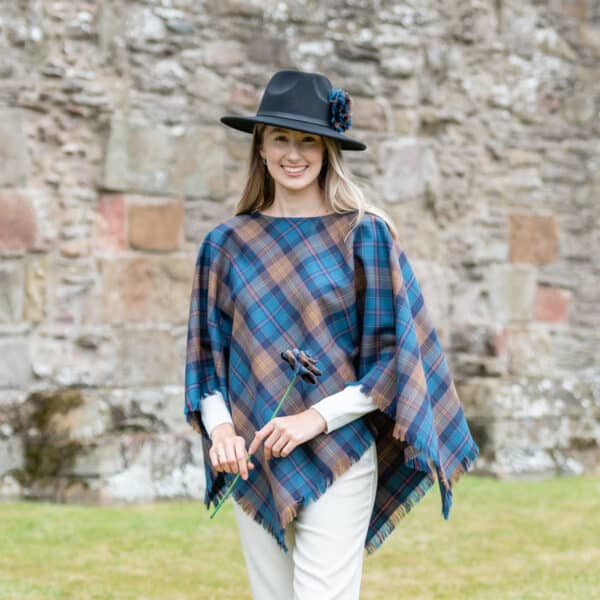 A woman in a Light Weight 11oz Premium Wool Tartan Poncho, standing in front of a stone wall.