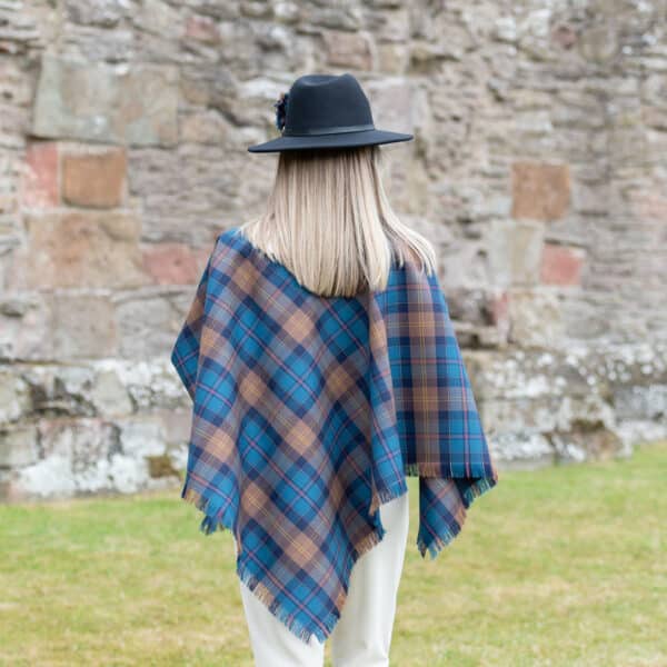 A woman wearing a Light Weight 11oz Premium Wool Tartan Poncho made from light weight 11oz premium wool fabric in front of a stone wall.