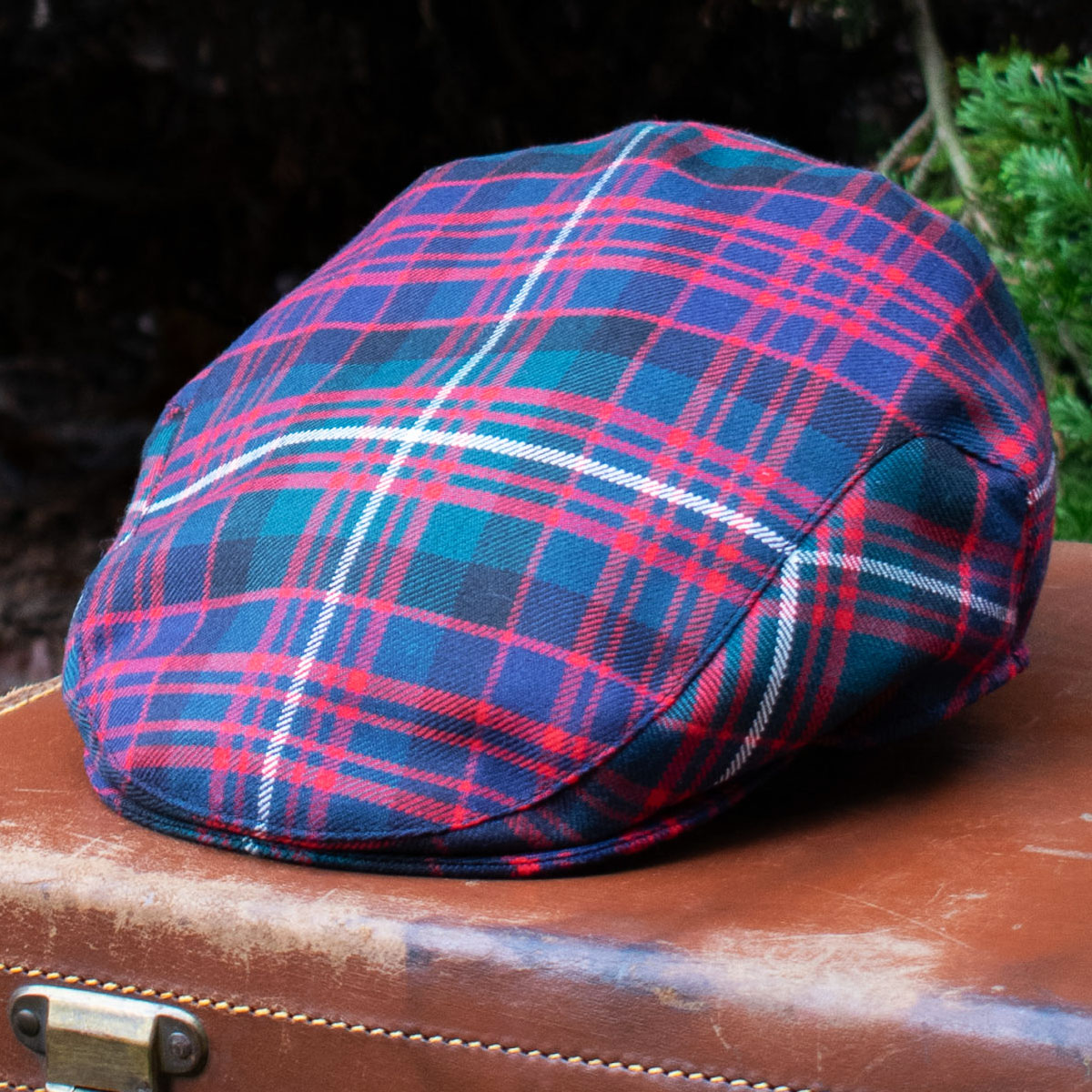 A Premium Wool Tartan Flat Cap in red, blue, and green rests on a closed brown leather suitcase outdoors.