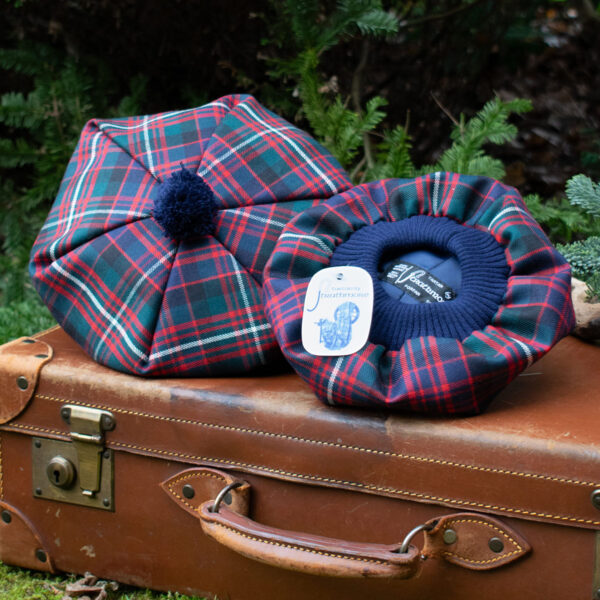 Two Premium Wool Tartan Tams with a pom-pom on top rest on a vintage brown leather suitcase. One tam is right side up, and the other is upside down, showing the brand label inside. Bushes are in the background.