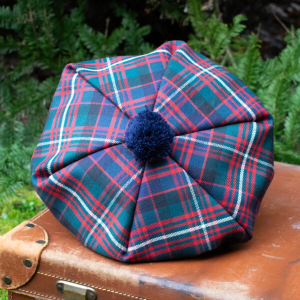 A Premium Wool Tartan Tam featuring red, green, and blue patterns with a blue pompom on top is placed on a leather suitcase against a backdrop of lush green foliage.