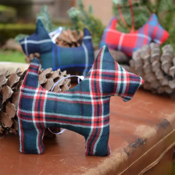 Tartan Scottie Dog Ornament, surrounded by pinecones, rests on a leather surface with a blurred outdoor backdrop.