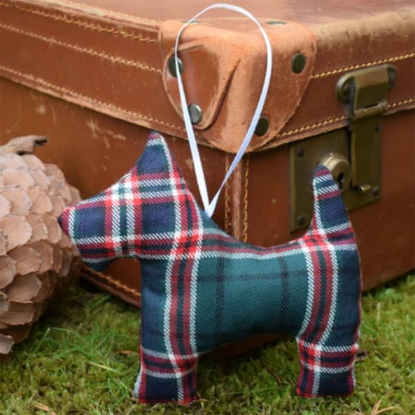 A Tartan Scottie Dog Ornament with a white ribbon for hanging is placed on green moss in front of a vintage brown suitcase.