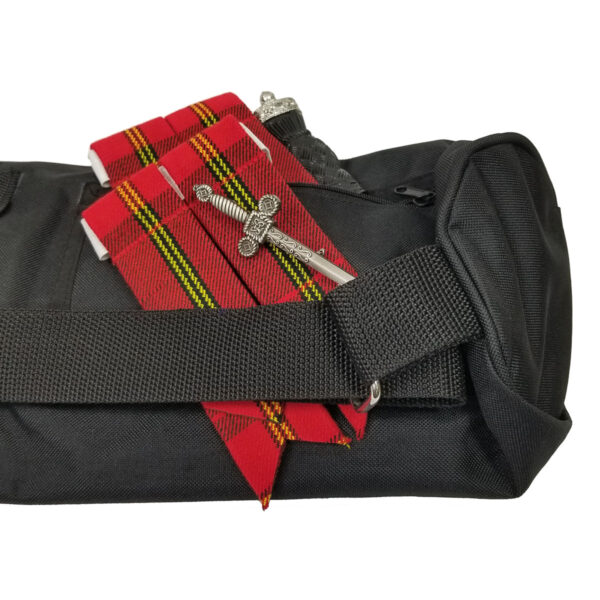 A black bag with a folded red, yellow, and black tartan-patterned sash, featuring a small ornamental silver sword, is in focus.