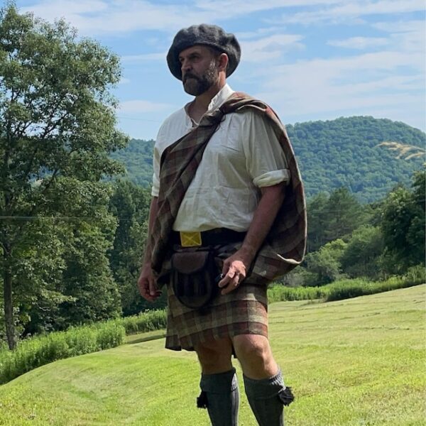 A person wearing traditional Scottish attire, including a kilt and beret, stands outdoors in a grassy area with trees and hills in the background.