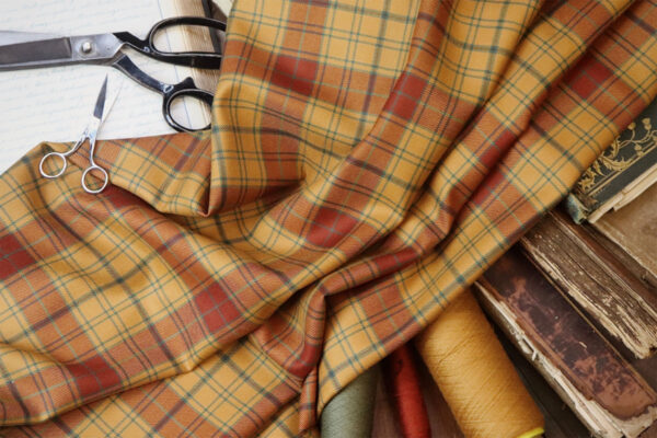 A piece of Glen Affric Tartan fabric is draped over several colored threads and books, with a pair of scissors and fabric shears placed on the side.