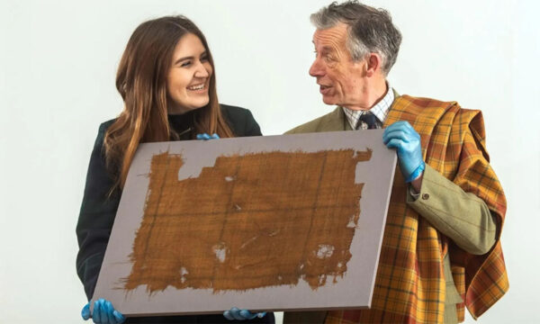 Two people are holding an aged piece of fabric mounted on a board. They are both wearing blue gloves and smiling while looking at each other, marveling at the exquisite Glen Affric Tartan by the Yard they have preserved.