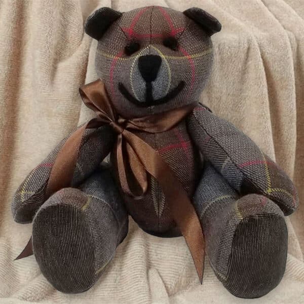 An Outlander Tartan Heritage Bears & Roses teddy bear, featuring a lightweight 11oz premium wool tartan design and a brown ribbon around its neck, is seated against a beige cloth backdrop.