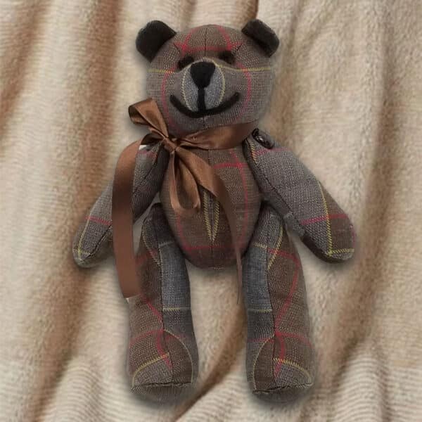 An 11oz lightweight, premium wool Outlander Tartan Heritage Bear adorned with a brown ribbon around its neck is set against a beige fabric background.