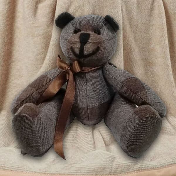 The Outlander Tartan Heritage Bears & Roses, a lightweight 11oz premium wool tartan bear adorned with a brown ribbon around its neck, is sitting against a beige fabric background.