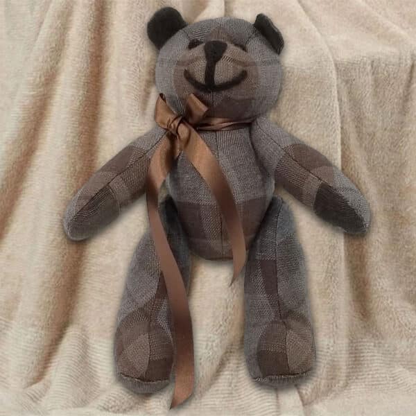 An 11oz lightweight premium wool tartan bear from the Outlander Tartan Heritage Bears & Roses collection, adorned with a brown ribbon, rests on a beige textured fabric background.