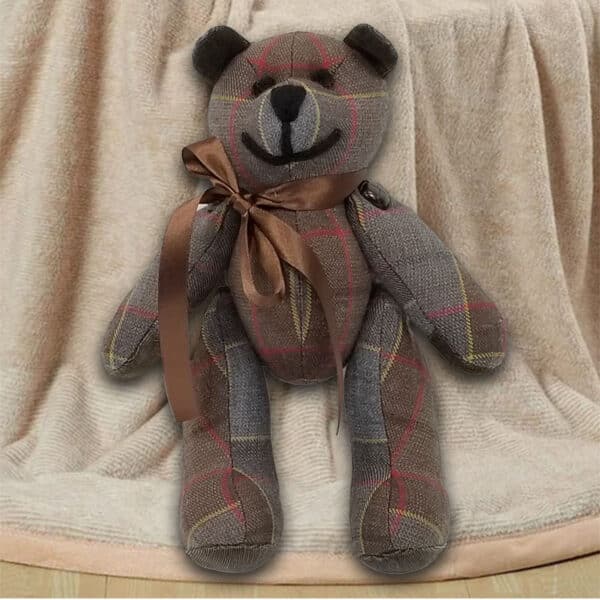 The Outlander Tartan Heritage Bears & Roses plush toy bear, adorned with a brown ribbon around its neck, is set against a beige-textured backdrop. Made from lightweight 11oz premium wool, this endearing bear showcases a delightful grey and red plaid pattern.