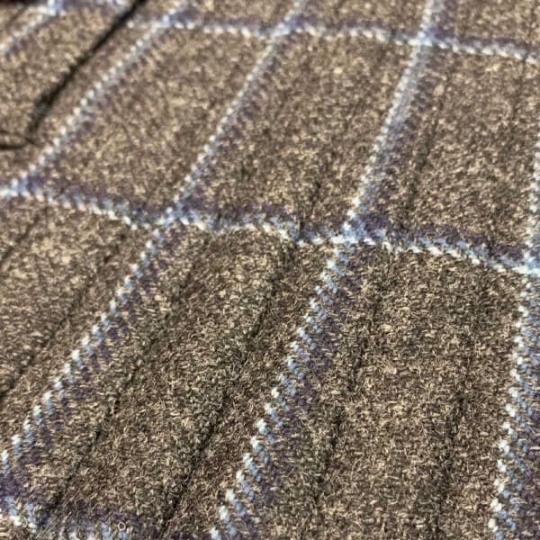 Close-up of a Twill Tweed Kilt showcasing a grey wool textile with a blue and white plaid pattern. The fabric appears textured and warm, reminiscent of traditional tweed.