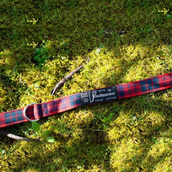 A Tartan Dog Leash made from 11oz lightweight premium wool, featuring a red and black pattern with a silver D-ring, rests on a grassy surface.